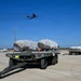 A military airfield scene showing two flatbed trailers loaded with large tarp-covered cargo secured with orange straps, positioned on a concrete apron. A large military aircraft, consistent with a B-52 Stratofortress, is visible flying overhead against a clear blue sky. A white utility vehicle and a light pole are visible in the background near a perimeter fence.