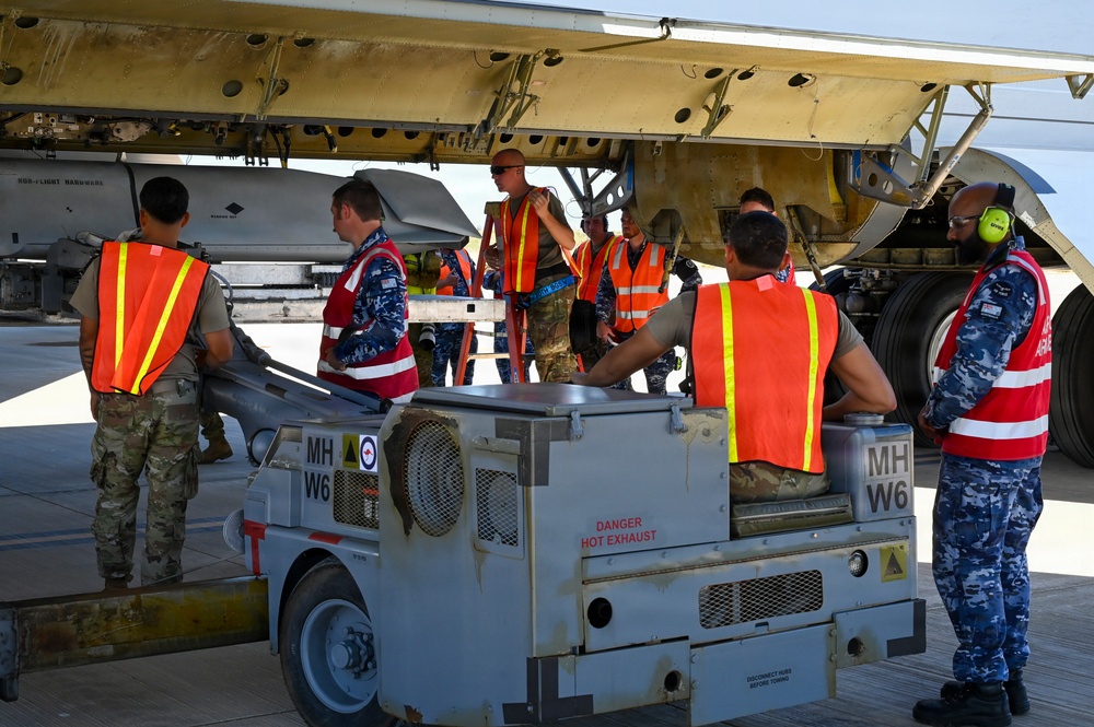 Military personnel from what appear to be multiple nations are conducting aircraft maintenance or weapons loading operations beneath the wing of a large military aircraft. Ground crew members wearing orange and red safety vests are gathered around ground support equipment marked 'MH W6' with a 'DANGER HOT EXHAUST' warning label. The scene takes place on a flight line or tarmac, suggesting a joint military exercise or operation.