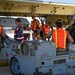 Military personnel from what appear to be multiple nations are conducting aircraft maintenance or weapons loading operations beneath the wing of a large military aircraft. Ground crew members wearing orange and red safety vests are gathered around ground support equipment marked 'MH W6' with a 'DANGER HOT EXHAUST' warning label. The scene takes place on a flight line or tarmac, suggesting a joint military exercise or operation.