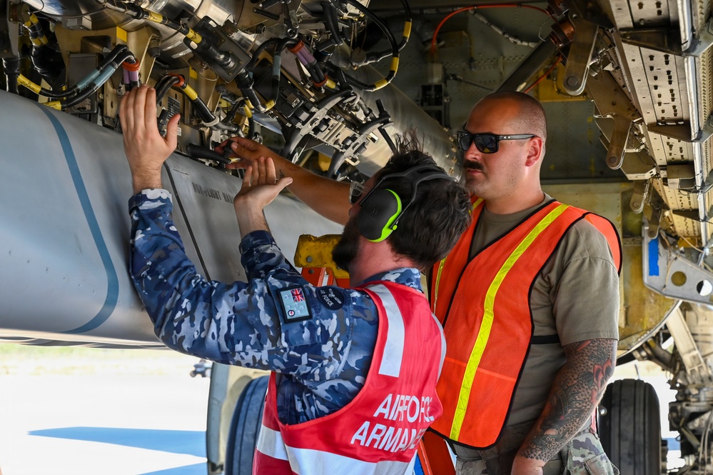 Three military personnel are working on the underside of a military aircraft, appearing to load or inspect an ordnance/munition. Two individuals wear safety vests — one red labeled 'AIRFORCE ARMAMENT' and one orange with reflective striping — while a third wears blue camouflage uniform with what appears to be a British/Australian flag patch. The scene depicts a joint multinational armament loading or maintenance operation.