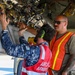 Three military personnel are working on the underside of a military aircraft, appearing to load or inspect an ordnance/munition. Two individuals wear safety vests — one red labeled 'AIRFORCE ARMAMENT' and one orange with reflective striping — while a third wears blue camouflage uniform with what appears to be a British/Australian flag patch. The scene depicts a joint multinational armament loading or maintenance operation.