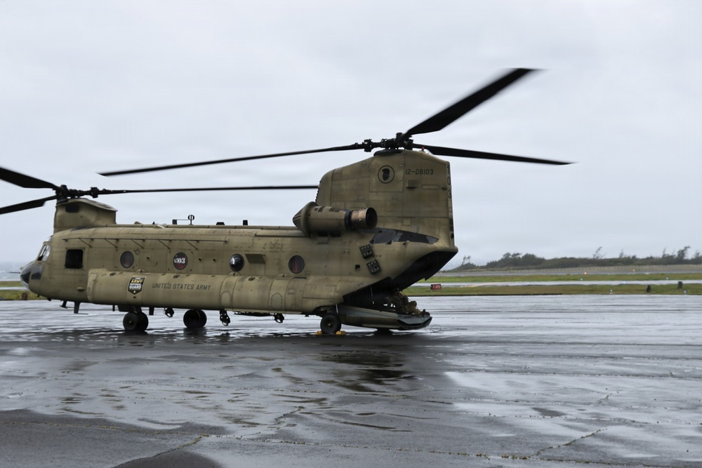 A United States Army CH-47 Chinook tandem-rotor helicopter is parked on a wet tarmac under overcast skies. The aircraft bears the serial number 12-08103 and displays Army National Guard markings. The rotors appear to be spinning or recently active, as evidenced by motion blur on the blades.