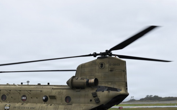 A United States Army CH-47 Chinook tandem-rotor helicopter is parked on a wet tarmac under overcast skies. The aircraft bears the serial number 12-08103 and displays Army National Guard markings. The rotors appear to be spinning or recently active, as evidenced by motion blur on the blades.
