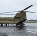 A United States Army CH-47 Chinook tandem-rotor helicopter is parked on a wet tarmac under overcast skies. The aircraft bears the serial number 12-08103 and displays Army National Guard markings. The rotors appear to be spinning or recently active, as evidenced by motion blur on the blades.