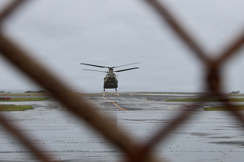 A tandem-rotor military helicopter, consistent with a CH-47 Chinook, is photographed through a chain-link fence on a wet airfield tarmac under overcast skies. The image is shot from a low angle through a gap in the fence, creating a framed perspective. The wet surface and gray sky suggest recent or ongoing rainfall.