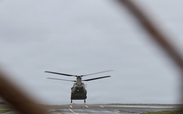 A tandem-rotor military helicopter, consistent with a CH-47 Chinook, is photographed through a chain-link fence on a wet airfield tarmac under overcast skies. The image is shot from a low angle through a gap in the fence, creating a framed perspective. The wet surface and gray sky suggest recent or ongoing rainfall.
