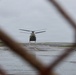 A tandem-rotor military helicopter, consistent with a CH-47 Chinook, is photographed through a chain-link fence on a wet airfield tarmac under overcast skies. The image is shot from a low angle through a gap in the fence, creating a framed perspective. The wet surface and gray sky suggest recent or ongoing rainfall.