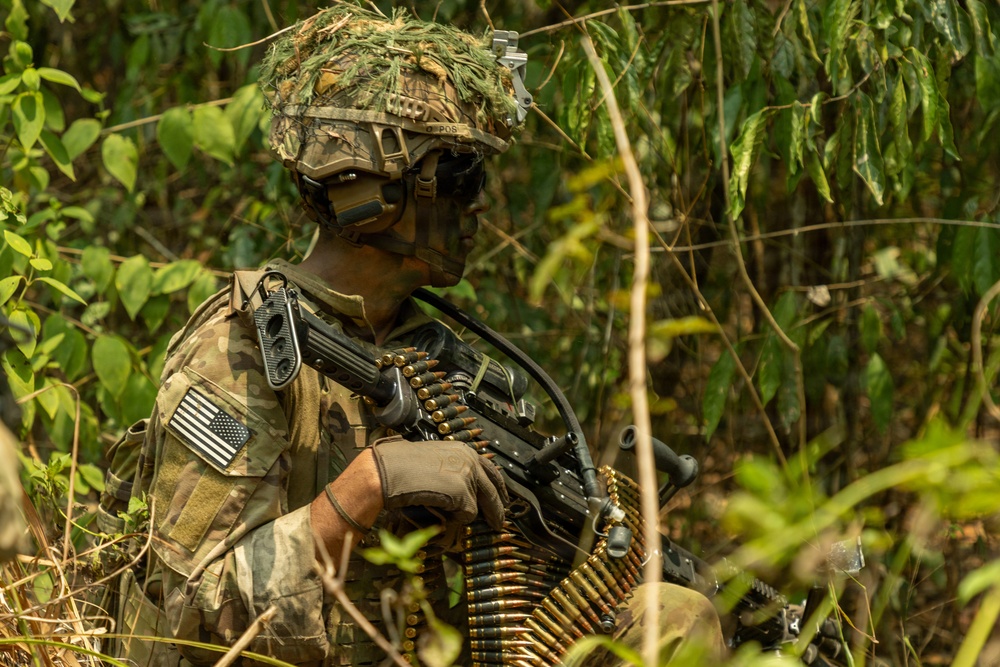 A U.S. Army soldier in full combat gear is positioned in a dense jungle or forested environment. The soldier is equipped with a machine gun and a belt of linked ammunition, wearing MultiCam camouflage with a ghillie-covered helmet. The image appears to document a military training exercise or operational patrol in a tropical setting.