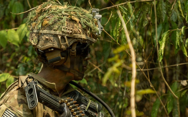 A U.S. Army soldier in full combat gear is positioned in a dense jungle or forested environment. The soldier is equipped with a machine gun and a belt of linked ammunition, wearing MultiCam camouflage with a ghillie-covered helmet. The image appears to document a military training exercise or operational patrol in a tropical setting.