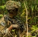 A U.S. Army soldier in full combat gear is positioned in a dense jungle or forested environment. The soldier is equipped with a machine gun and a belt of linked ammunition, wearing MultiCam camouflage with a ghillie-covered helmet. The image appears to document a military training exercise or operational patrol in a tropical setting.
