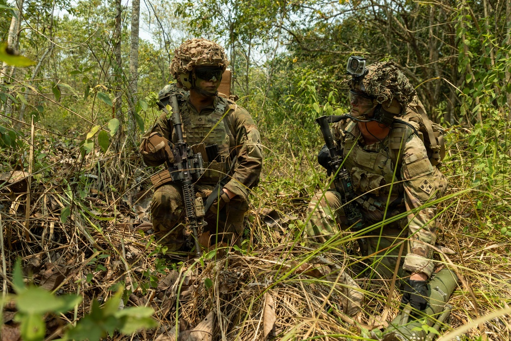 Two military personnel in full combat gear are positioned in a dense jungle or forest environment, appearing to be in a tactical crouch or kneeling position. Both soldiers are equipped with camouflage helmets, body armor, and assault rifles, suggesting a training exercise or patrol operation. The lush, overgrown vegetation and dry leaf ground cover indicate a tropical or subtropical environment.