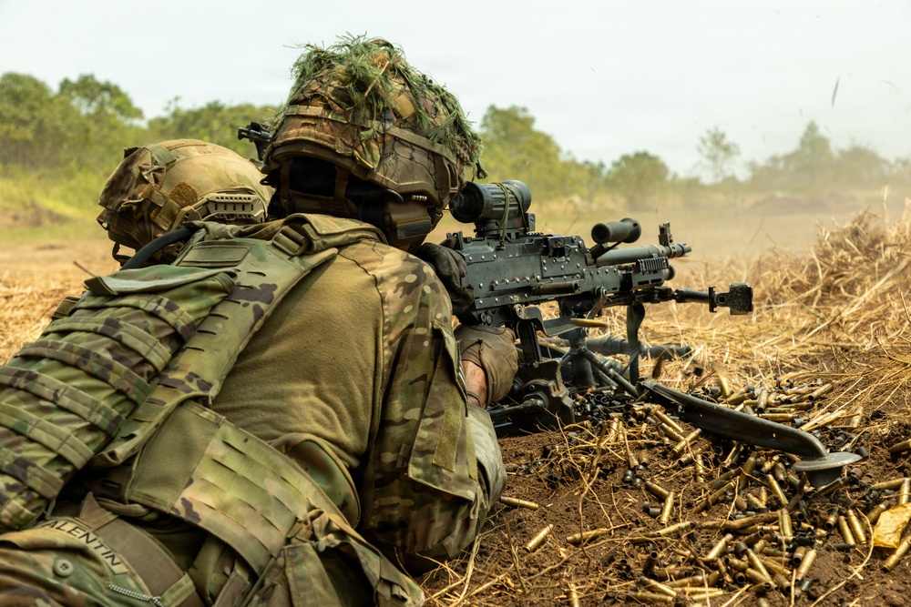 Two soldiers in full combat gear are operating a crew-served machine gun from a prone position in an open field. A large number of expended brass casings are scattered around the weapon, indicating sustained fire has taken place. The soldiers are wearing MultiCam/OCP camouflage uniforms with tactical helmets, one adorned with ghillie-style foliage.