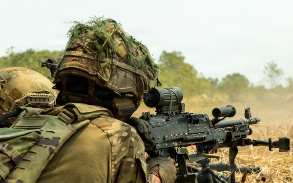 Two soldiers in full combat gear are operating a crew-served machine gun from a prone position in an open field. A large number of expended brass casings are scattered around the weapon, indicating sustained fire has taken place. The soldiers are wearing MultiCam/OCP camouflage uniforms with tactical helmets, one adorned with ghillie-style foliage.
