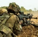 Two soldiers in full combat gear are operating a crew-served machine gun from a prone position in an open field. A large number of expended brass casings are scattered around the weapon, indicating sustained fire has taken place. The soldiers are wearing MultiCam/OCP camouflage uniforms with tactical helmets, one adorned with ghillie-style foliage.