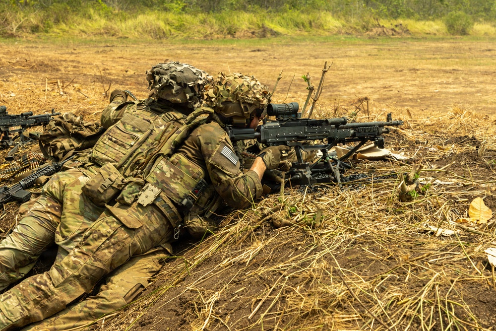 Two soldiers in MultiCam camouflage uniforms are in a prone firing position on dry, open terrain. The forward soldier is aiming what appears to be an M240 machine gun mounted on a bipod, while the rear soldier provides support. Both wear helmets with camouflage covers and full tactical gear including plate carriers and pouches.