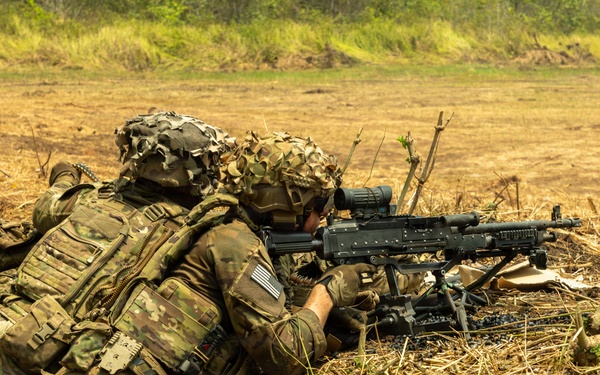 Two soldiers in MultiCam camouflage uniforms are in a prone firing position on dry, open terrain. The forward soldier is aiming what appears to be an M240 machine gun mounted on a bipod, while the rear soldier provides support. Both wear helmets with camouflage covers and full tactical gear including plate carriers and pouches.
