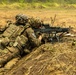 Two soldiers in MultiCam camouflage uniforms are in a prone firing position on dry, open terrain. The forward soldier is aiming what appears to be an M240 machine gun mounted on a bipod, while the rear soldier provides support. Both wear helmets with camouflage covers and full tactical gear including plate carriers and pouches.