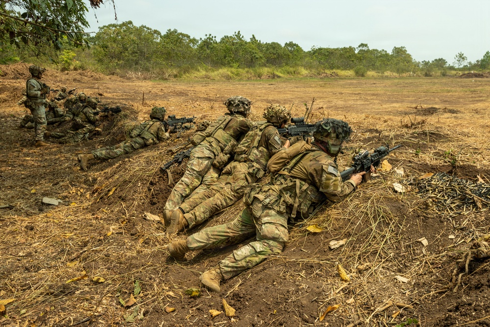 The image shows multiple soldiers in prone firing positions along a shallow trench or berm in an open field during what appears to be a military training exercise. The soldiers are wearing MultiCam (OCP) camouflage uniforms with full combat gear and are aiming rifles downrange. Spent brass casings are visible on the ground to the right, indicating live or blank fire has occurred.