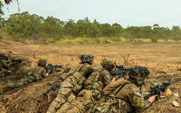 The image shows multiple soldiers in prone firing positions along a shallow trench or berm in an open field during what appears to be a military training exercise. The soldiers are wearing MultiCam (OCP) camouflage uniforms with full combat gear and are aiming rifles downrange. Spent brass casings are visible on the ground to the right, indicating live or blank fire has occurred.