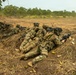 The image shows multiple soldiers in prone firing positions along a shallow trench or berm in an open field during what appears to be a military training exercise. The soldiers are wearing MultiCam (OCP) camouflage uniforms with full combat gear and are aiming rifles downrange. Spent brass casings are visible on the ground to the right, indicating live or blank fire has occurred.