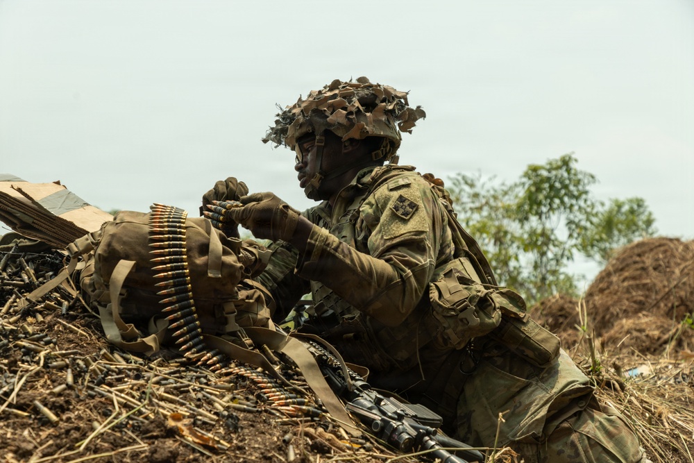 A soldier in full camouflage gear is positioned behind an earthen berm or fighting position, handling a belt of ammunition. The soldier is wearing a helmet with camouflage netting and foliage attached, along with tactical body armor and gloves. The setting appears to be an outdoor training exercise or operational environment with sparse vegetation and overcast skies.