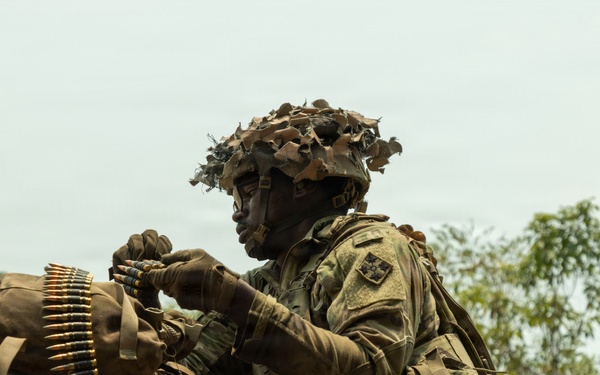 A soldier in full camouflage gear is positioned behind an earthen berm or fighting position, handling a belt of ammunition. The soldier is wearing a helmet with camouflage netting and foliage attached, along with tactical body armor and gloves. The setting appears to be an outdoor training exercise or operational environment with sparse vegetation and overcast skies.
