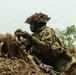 A soldier in full camouflage gear is positioned behind an earthen berm or fighting position, handling a belt of ammunition. The soldier is wearing a helmet with camouflage netting and foliage attached, along with tactical body armor and gloves. The setting appears to be an outdoor training exercise or operational environment with sparse vegetation and overcast skies.