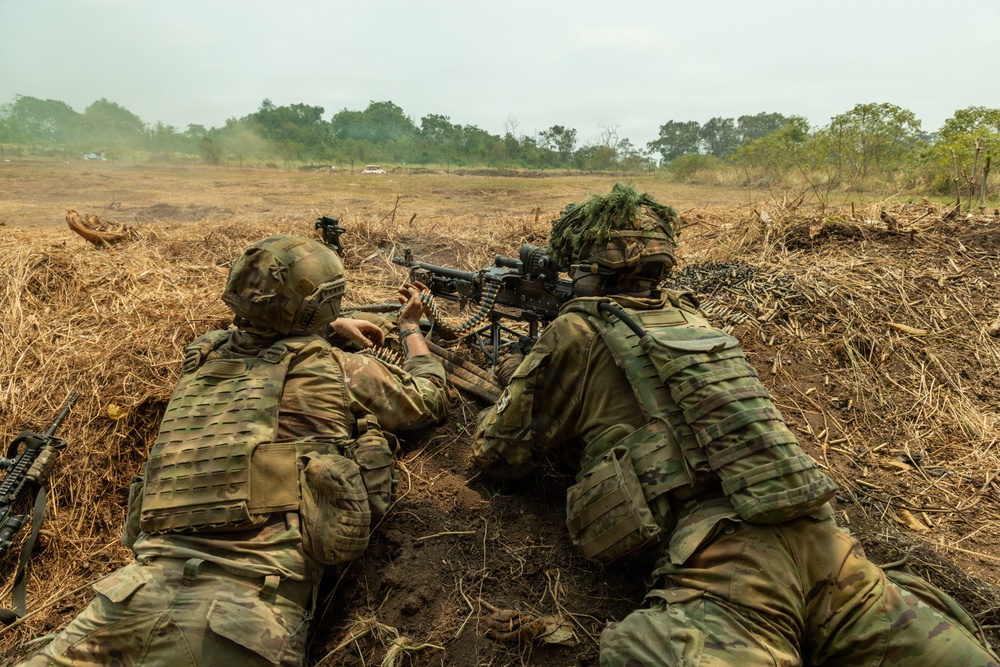 Two soldiers in multicam camouflage uniforms are positioned in a shallow fighting position or trench, operating a belt-fed machine gun in an open field environment. The gunner on the right is aiming downrange while the soldier on the left appears to be managing the ammunition belt. The terrain is dry and open with sparse vegetation and trees visible in the background.