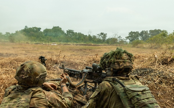 Two soldiers in multicam camouflage uniforms are positioned in a shallow fighting position or trench, operating a belt-fed machine gun in an open field environment. The gunner on the right is aiming downrange while the soldier on the left appears to be managing the ammunition belt. The terrain is dry and open with sparse vegetation and trees visible in the background.