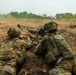 Two soldiers in multicam camouflage uniforms are positioned in a shallow fighting position or trench, operating a belt-fed machine gun in an open field environment. The gunner on the right is aiming downrange while the soldier on the left appears to be managing the ammunition belt. The terrain is dry and open with sparse vegetation and trees visible in the background.