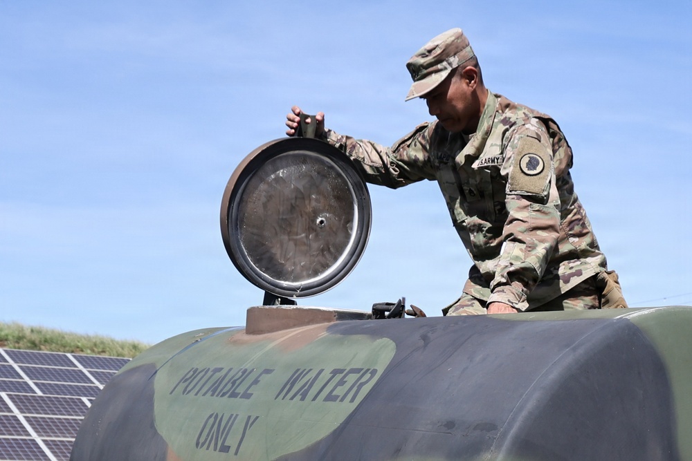 A U.S. Army soldier in OCP (Operational Camouflage Pattern) uniform is opening the hatch of a military potable water tank. The tank is painted olive drab and labeled 'POTABLE WATER ONLY' in stenciled lettering. Solar panels are visible in the background, suggesting a forward operating base or installation with renewable energy infrastructure.