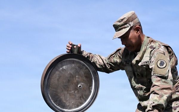 A U.S. Army soldier in OCP (Operational Camouflage Pattern) uniform is opening the hatch of a military potable water tank. The tank is painted olive drab and labeled 'POTABLE WATER ONLY' in stenciled lettering. Solar panels are visible in the background, suggesting a forward operating base or installation with renewable energy infrastructure.