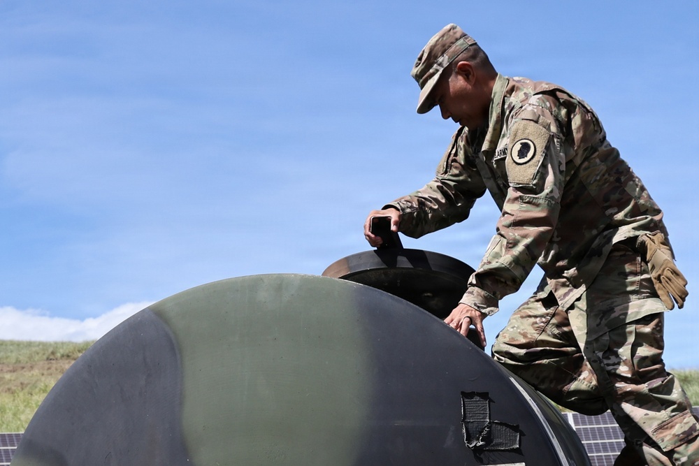 A U.S. Army soldier in Operational Camouflage Pattern (OCP) uniform is working on a large dark green dome-shaped structure, possibly a water storage tank or equipment housing. The soldier appears to be inspecting or operating a hatch or access point on top of the curved surface. Solar panels are partially visible in the lower right corner of the image.