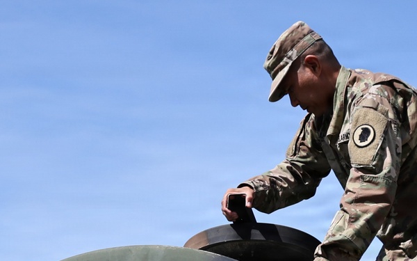 A U.S. Army soldier in Operational Camouflage Pattern (OCP) uniform is working on a large dark green dome-shaped structure, possibly a water storage tank or equipment housing. The soldier appears to be inspecting or operating a hatch or access point on top of the curved surface. Solar panels are partially visible in the lower right corner of the image.