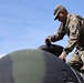 A U.S. Army soldier in Operational Camouflage Pattern (OCP) uniform is working on a large dark green dome-shaped structure, possibly a water storage tank or equipment housing. The soldier appears to be inspecting or operating a hatch or access point on top of the curved surface. Solar panels are partially visible in the lower right corner of the image.