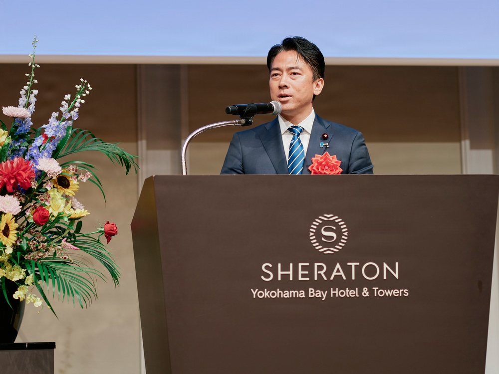 A man in a dark blue suit with a striped tie is delivering a speech at a podium inside the Sheraton Yokohama Bay Hotel &amp; Towers. He is wearing a lapel pin resembling a Japanese national flag badge and a red floral corsage on his chest. A colorful floral arrangement is visible to his left.