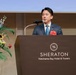 A man in a dark blue suit with a striped tie is delivering a speech at a podium inside the Sheraton Yokohama Bay Hotel &amp; Towers. He is wearing a lapel pin resembling a Japanese national flag badge and a red floral corsage on his chest. A colorful floral arrangement is visible to his left.