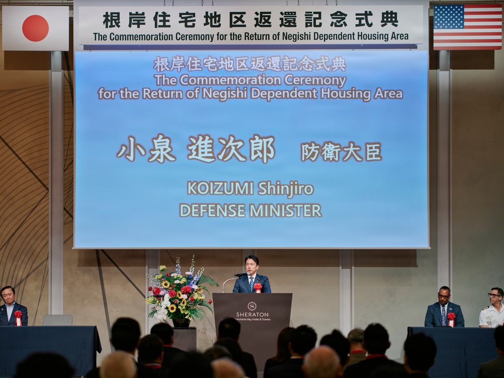 This image shows a formal commemoration ceremony for the return of the Negishi Dependent Housing Area, held at the Sheraton Yokohama Bay Hotel &amp; Towers. Japanese Defense Minister Koizumi Shinjiro is identified on the large projection screen as the speaker at the podium. The event features both Japanese and American flags, indicating a bilateral diplomatic occasion between Japan and the United States.