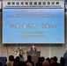 This image shows a formal commemoration ceremony for the return of the Negishi Dependent Housing Area, held at the Sheraton Yokohama Bay Hotel &amp; Towers. Japanese Defense Minister Koizumi Shinjiro is identified on the large projection screen as the speaker at the podium. The event features both Japanese and American flags, indicating a bilateral diplomatic occasion between Japan and the United States.