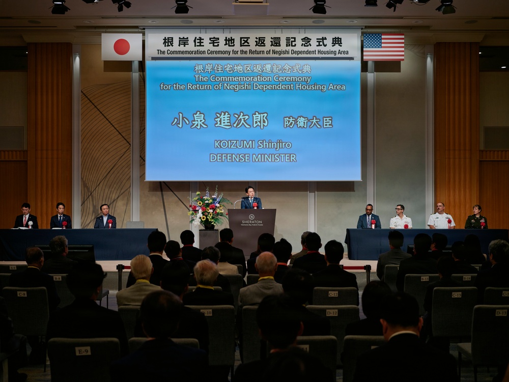 This is a formal commemoration ceremony for the return of the Negishi Dependent Housing Area, held at a Sheraton hotel venue. Japanese Defense Minister Koizumi Shinjiro is speaking at the podium, flanked by Japanese and American officials seated at a head table. Both Japanese and American flags are displayed, indicating a bilateral diplomatic event.