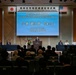 This is a formal commemoration ceremony for the return of the Negishi Dependent Housing Area, held at a Sheraton hotel venue. Japanese Defense Minister Koizumi Shinjiro is speaking at the podium, flanked by Japanese and American officials seated at a head table. Both Japanese and American flags are displayed, indicating a bilateral diplomatic event.