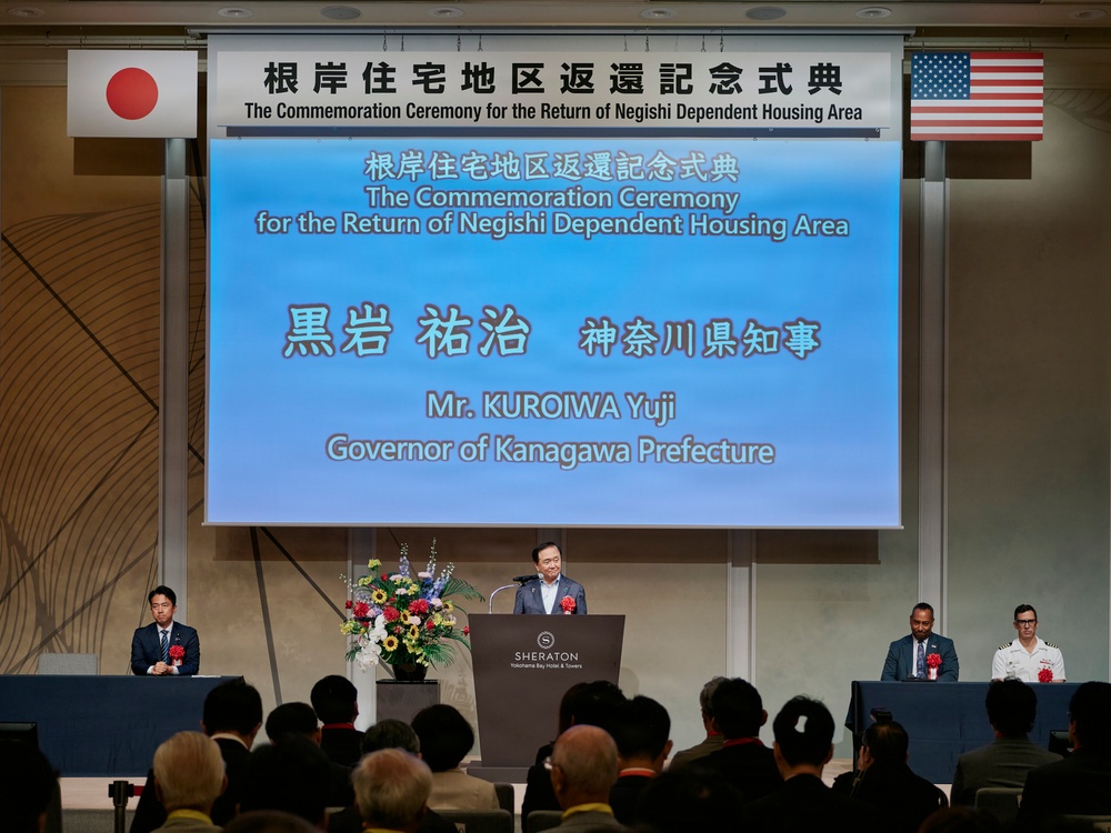 This image shows a formal commemoration ceremony for the return of the Negishi Dependent Housing Area, held at the Sheraton Yokohama Bay Hotel &amp; Towers. Mr. Kuroiwa Yuji, Governor of Kanagawa Prefecture, is delivering a speech at the podium. The event features both Japanese and American flags, indicating a bilateral Japan-U.S. occasion.
