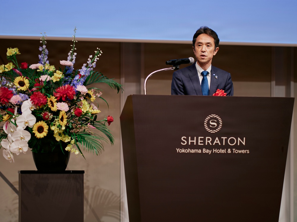 A man in a dark suit with a blue tie is delivering a speech at a podium bearing the Sheraton Yokohama Bay Hotel &amp; Towers logo. He has a red boutonniere on his lapel, suggesting a formal or celebratory event. A large, colorful floral arrangement is displayed on a pedestal to his left.