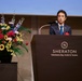 A man in a dark suit with a blue tie is delivering a speech at a podium bearing the Sheraton Yokohama Bay Hotel &amp; Towers logo. He has a red boutonniere on his lapel, suggesting a formal or celebratory event. A large, colorful floral arrangement is displayed on a pedestal to his left.