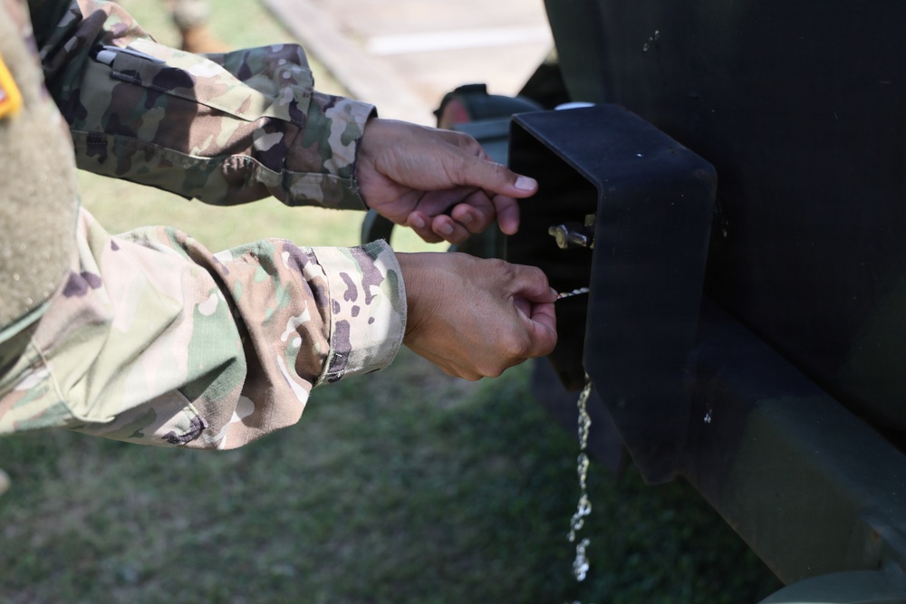 Two individuals wearing US Army OCP (Operational Camouflage Pattern) uniforms are working with their hands on a black metal bracket or mounting hardware attached to a dark-colored military vehicle or equipment. Water droplets are visibly dripping from the hardware, suggesting a fluid connection or fitting is being adjusted. The close-up perspective focuses entirely on the hands and the mechanical component being manipulated.