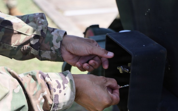 Two individuals wearing US Army OCP (Operational Camouflage Pattern) uniforms are working with their hands on a black metal bracket or mounting hardware attached to a dark-colored military vehicle or equipment. Water droplets are visibly dripping from the hardware, suggesting a fluid connection or fitting is being adjusted. The close-up perspective focuses entirely on the hands and the mechanical component being manipulated.