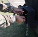 Two individuals wearing US Army OCP (Operational Camouflage Pattern) uniforms are working with their hands on a black metal bracket or mounting hardware attached to a dark-colored military vehicle or equipment. Water droplets are visibly dripping from the hardware, suggesting a fluid connection or fitting is being adjusted. The close-up perspective focuses entirely on the hands and the mechanical component being manipulated.