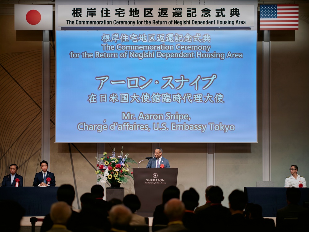 A formal commemoration ceremony is being held at the Sheraton Yokohama Bay Hotel &amp; Towers for the return of the Negishi Dependent Housing Area. Mr. Aaron Snipe, Chargé d'affaires of the U.S. Embassy Tokyo, is delivering a speech at the podium. The event features both Japanese and American flags, indicating a bilateral diplomatic occasion.