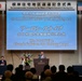 A formal commemoration ceremony is being held at the Sheraton Yokohama Bay Hotel &amp; Towers for the return of the Negishi Dependent Housing Area. Mr. Aaron Snipe, Chargé d'affaires of the U.S. Embassy Tokyo, is delivering a speech at the podium. The event features both Japanese and American flags, indicating a bilateral diplomatic occasion.