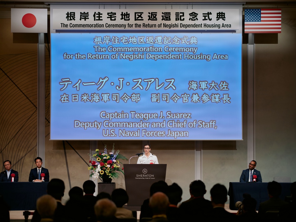 A formal commemoration ceremony is taking place at the Sheraton Yokohama Bay Hotel &amp; Towers for the return of the Negishi Dependent Housing Area. Captain Teague J. Suarez, Deputy Commander and Chief of Staff of U.S. Naval Forces Japan, is delivering a speech at the podium. The event features both Japanese and American flags, bilingual signage, and an audience of attendees seated in rows.